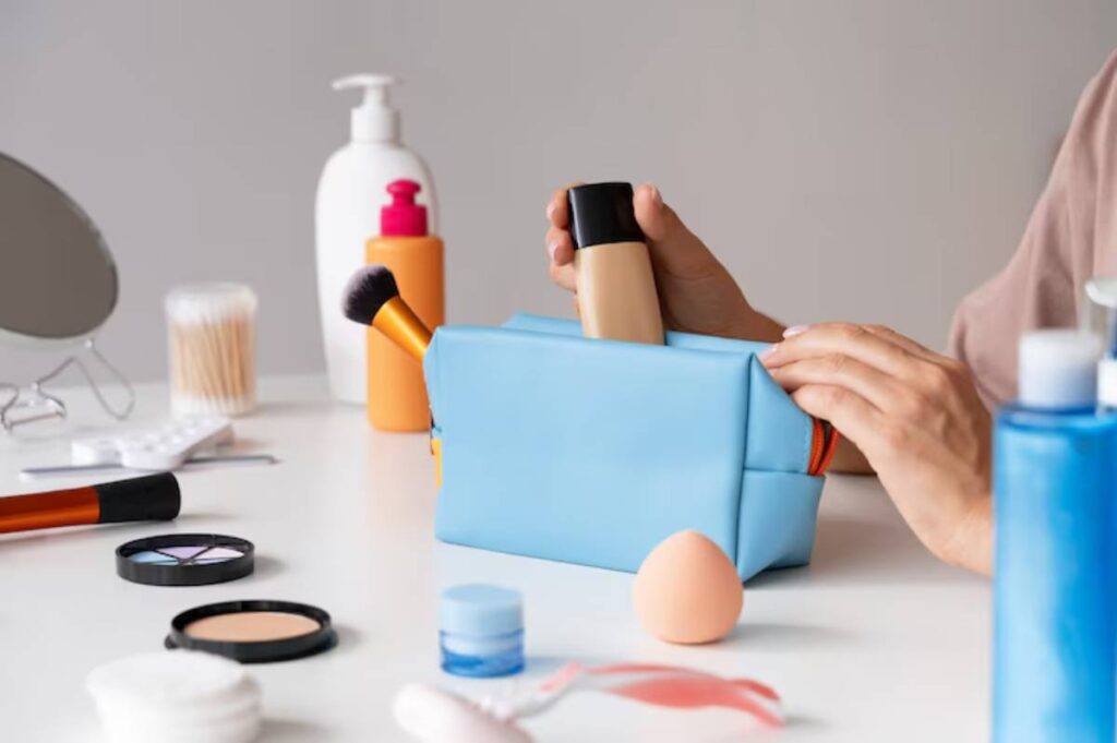 A hand reaches into a blue makeup bag, surrounded by various beauty products like foundation, brushes, and skincare items on a table.