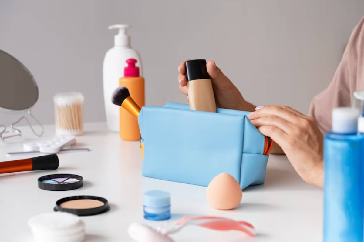 A hand reaches into a blue makeup bag, surrounded by various beauty products like foundation, brushes, and skincare items on a table.