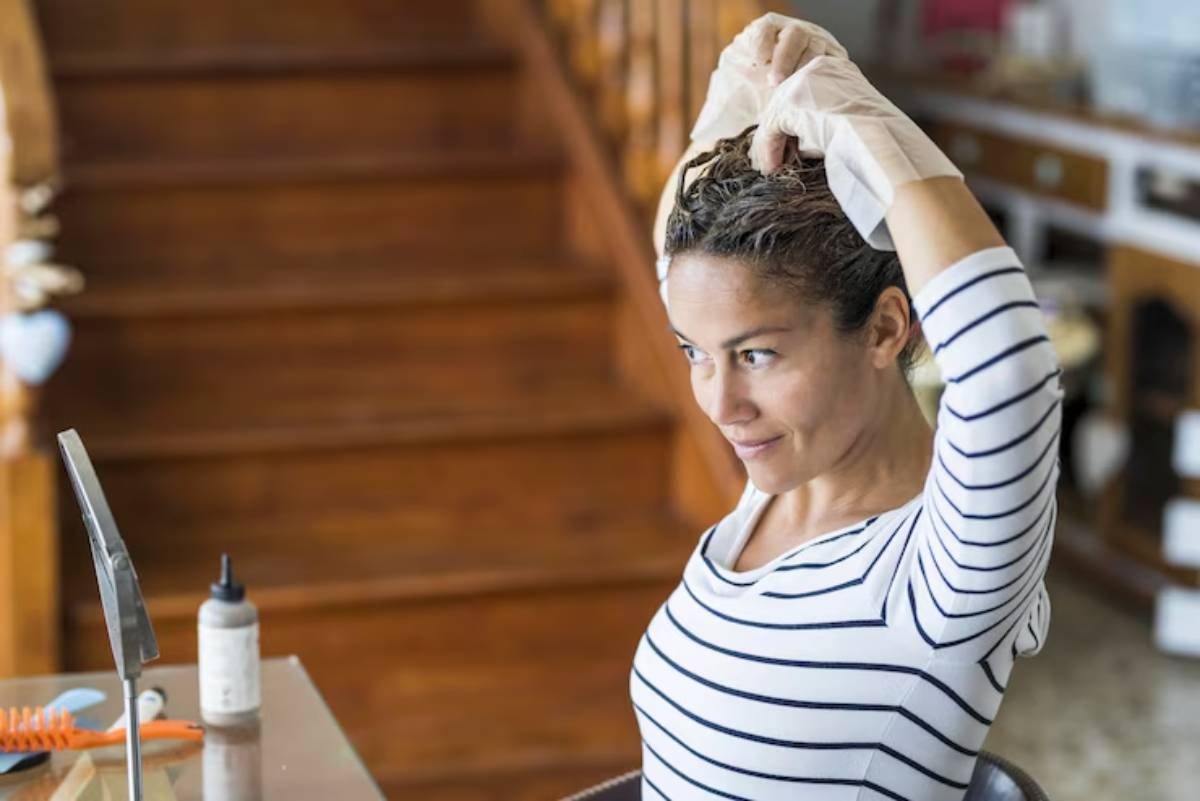 A woman in a striped shirt, wearing gloves, styles her hair in front of a mirror, with wooden stairs in the background.
