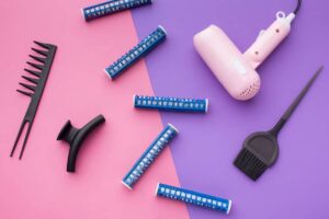 A pink hairdryer, blue hair rollers, a black comb, a styling brush, and a parting clip on a pink and purple background.