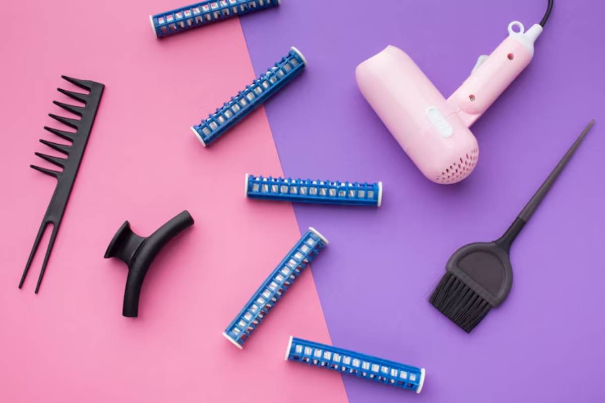 A pink hairdryer, blue hair rollers, a black comb, a styling brush, and a parting clip on a pink and purple background.