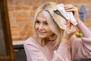 A woman applies hair dye using a brush while holding a piece of foil close to her hair against a rustic brick background.