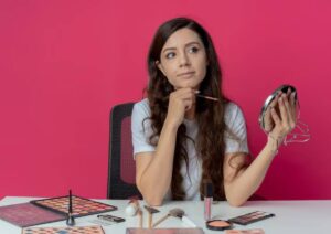 A woman holds a mirror and makeup brush, surrounded by various makeup products on a white table against a vibrant pink background.