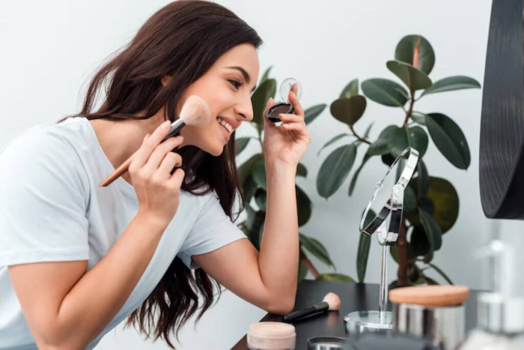 A person applies makeup with a brush while looking into a handheld mirror; makeup products and a plant are visible on the table.