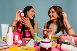 Two women showcase various beauty products, including makeup and skincare items, against a pastel blue background.