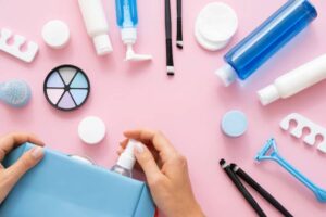 A person’s hand reaches for a spray bottle from a blue bag surrounded by various skincare products and tools on a pink background.