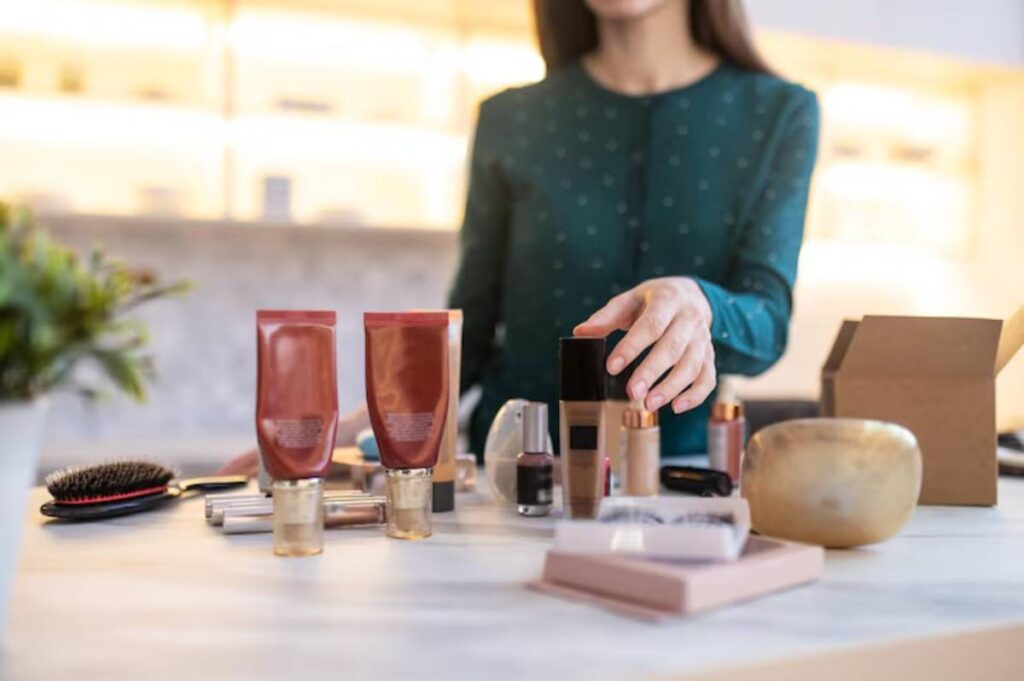 A woman in a green blouse arranges various makeup products and brushes on a table, alongside an open cardboard box.