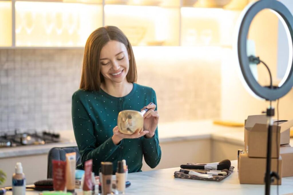 A woman in a green dotted top holds a skincare product, surrounded by makeup items and a ring light on a countertop in a bright kitchen.