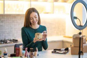 A woman in a green dotted top holds a skincare product, surrounded by makeup items and a ring light on a countertop in a bright kitchen.