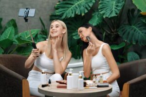 Two women in white outfits take a selfie while surrounded by skincare products on a table, with lush greenery in the background.