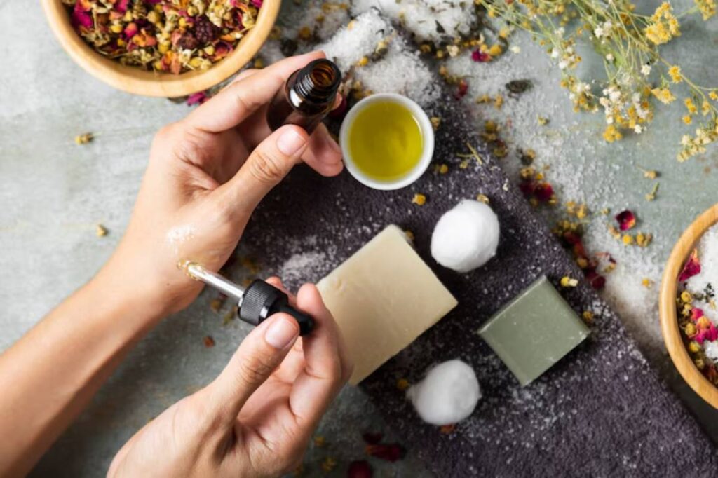 A person applies essential oil from a dropper to their wrist, surrounded by herbal ingredients, soap, and cotton balls.