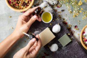 A person applies essential oil from a dropper to their wrist, surrounded by herbal ingredients, soap, and cotton balls.
