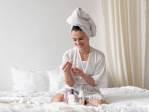 A person in a white robe and towel sits on a bed surrounded by various skincare products, preparing for a self-care routine.