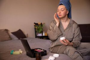 A person in a gray pajama set applies cream while sitting on a bed, surrounded by skincare products and a makeup palette.