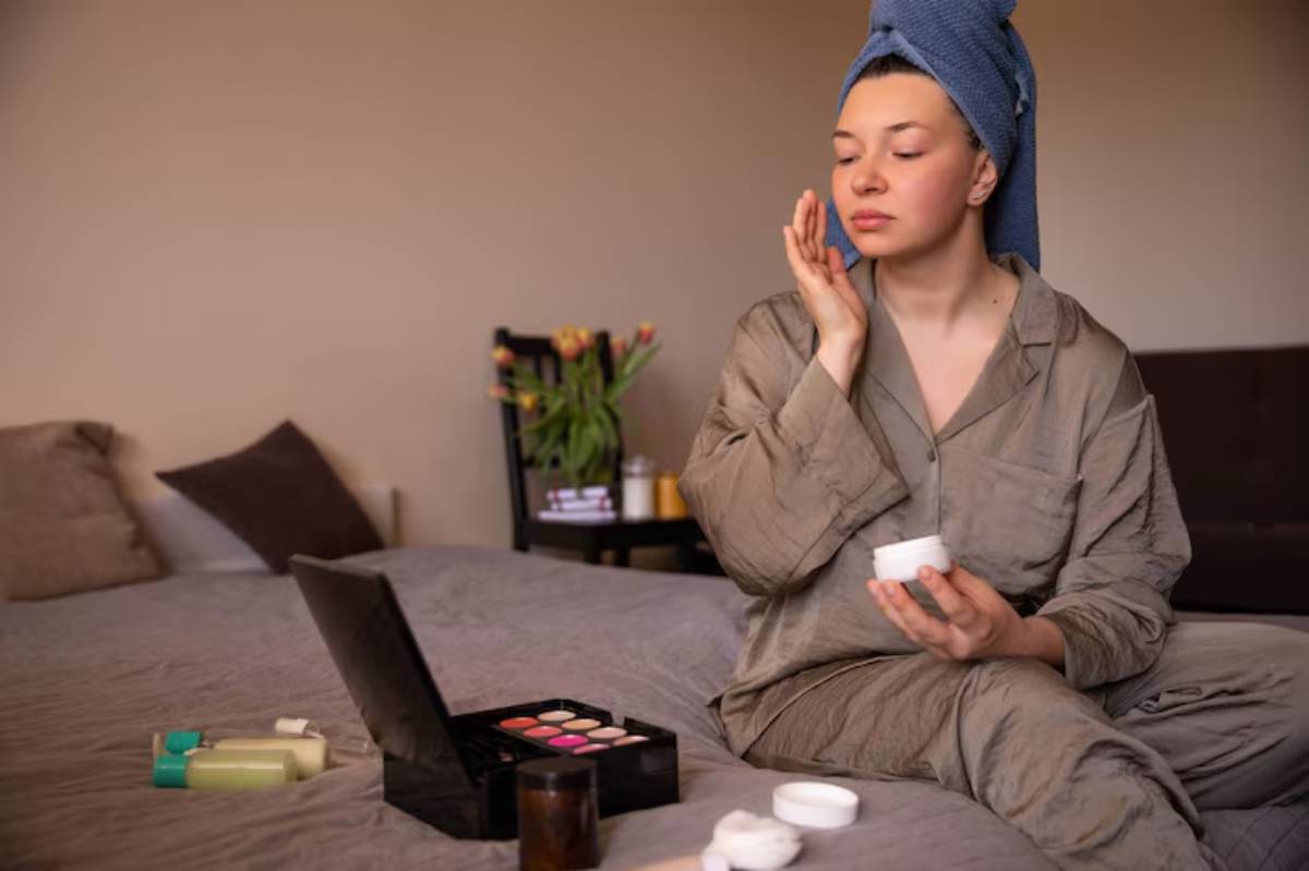 A person in a gray pajama set applies cream while sitting on a bed, surrounded by skincare products and a makeup palette.