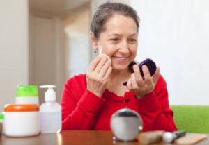 A person in a red sweater applies makeup using a compact mirror, surrounded by various beauty products on a wooden table.