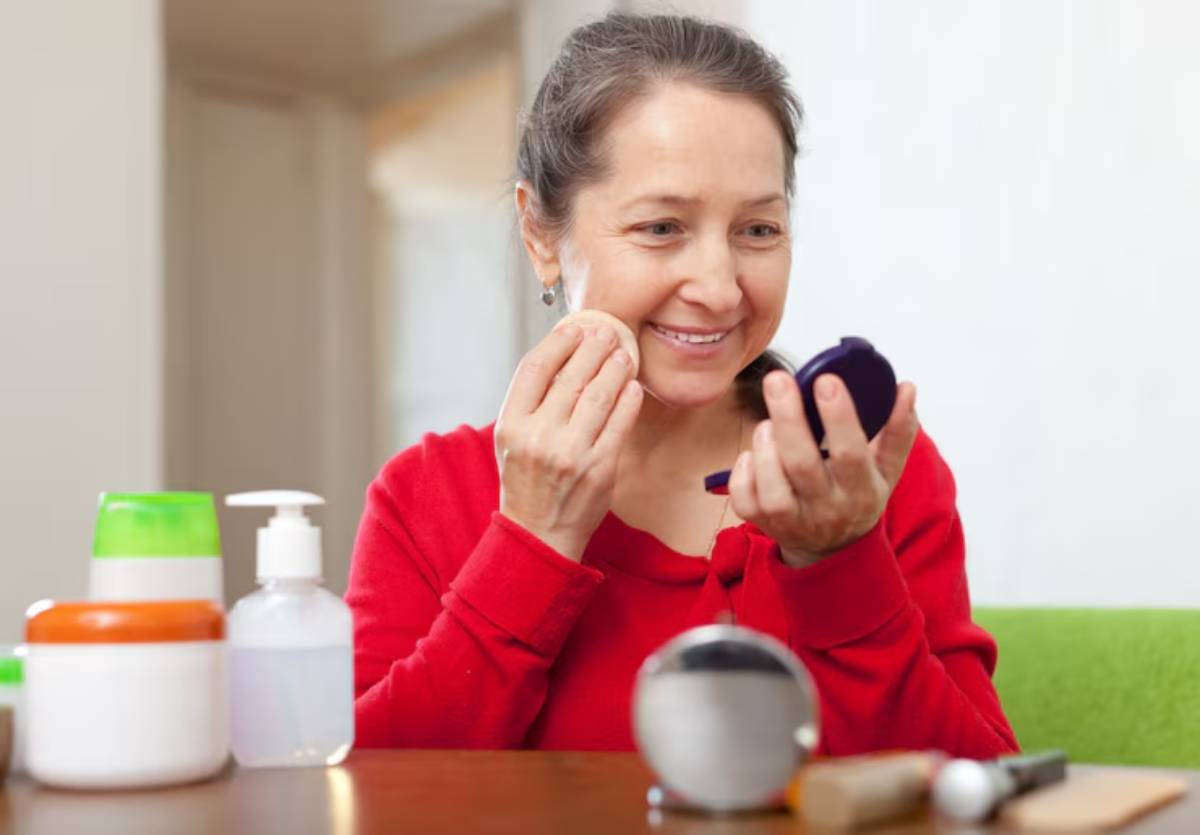 A person in a red sweater applies makeup using a compact mirror, surrounded by various beauty products on a wooden table.